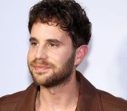 Ben Platt, wearing a brown jacket, smiles infront of a white background.