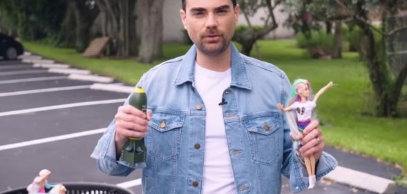 Ben Shapiro holds a Barbie Doll and a miniature replica bomb in the middle of a field.