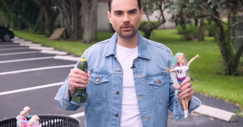 Ben Shapiro holds a Barbie Doll and a miniature replica bomb in the middle of a field.