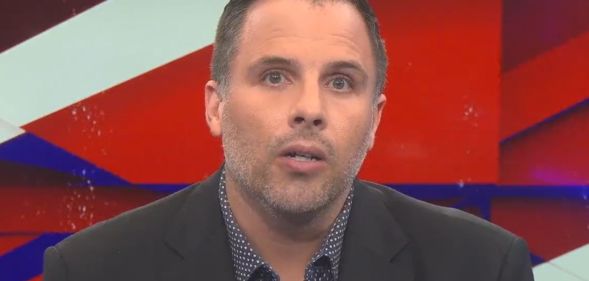 Dan Wootton, on the set of GB News, speaks towards a camera while wearing a suit and open collar shirt.