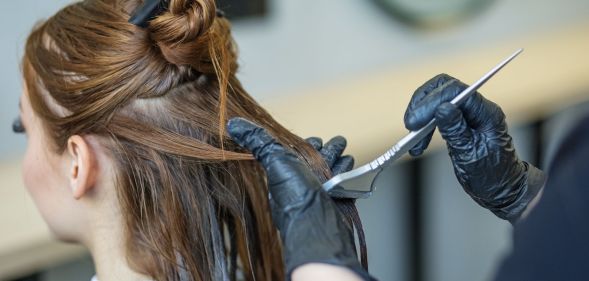 A person with brunette hair sits as someone with a hair dye brush, wearing black gloves, wipes dye on the back of their hair.