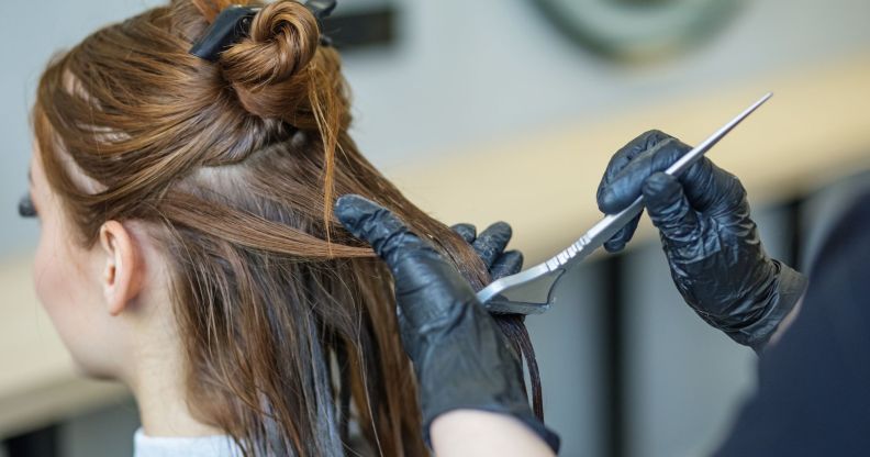 A person with brunette hair sits as someone with a hair dye brush, wearing black gloves, wipes dye on the back of their hair.