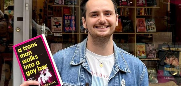 A picture of Harry Nicholas, in a denim jacket, holding a book that reads "A Trans Man Walks into a Gay Bar."