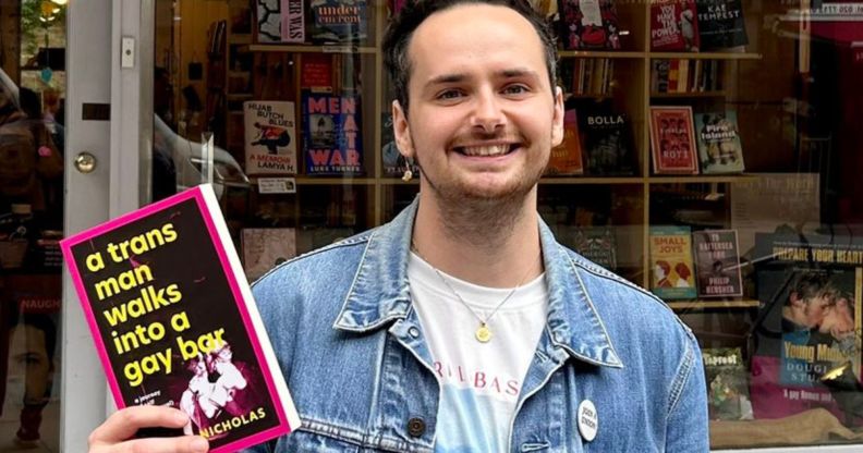 A picture of Harry Nicholas, in a denim jacket, holding a book that reads "A Trans Man Walks into a Gay Bar."