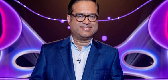 Paul Sinha in a blue suit and shirt standing in front of a blue and purple background.