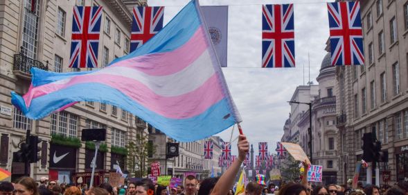A person holds a trans flag in the air at a crowded protest, with four union jacks being hung across two buildings.