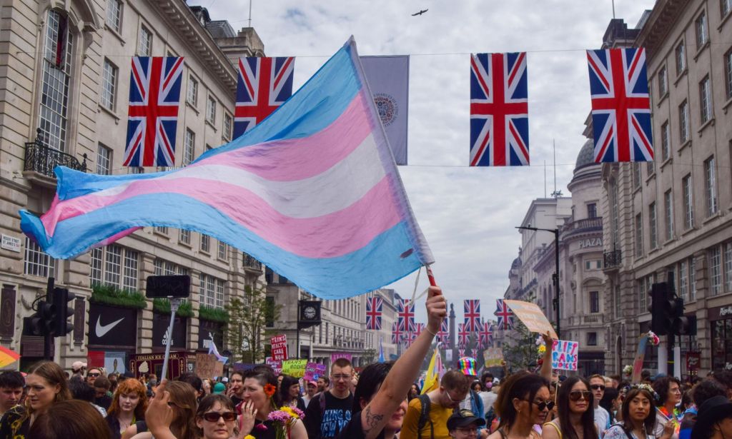 A person holds a trans flag in the air at a crowded protest, with four union jacks being hung across two buildings.