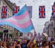 A person holds a trans flag in the air at a crowded protest, with four union jacks being hung across two buildings.