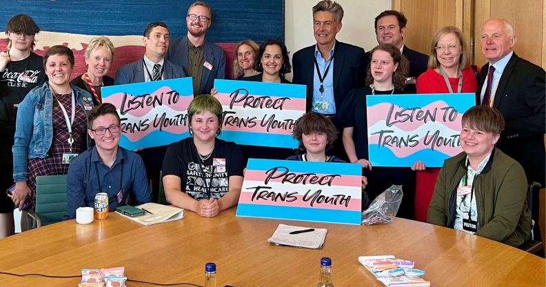 A group of MPs and trans youth sit around a wooden table, holding signs that say "protect trans youth" and "listen to trans youth."