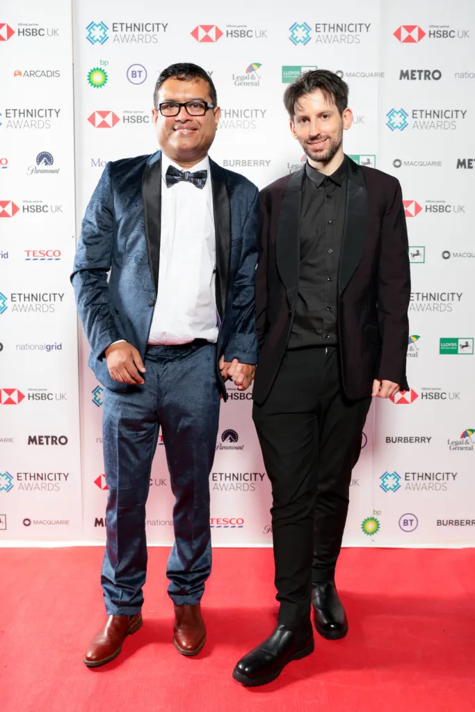 Paul Sinha and his partner Oliver Levy wear suits on a red carpet.