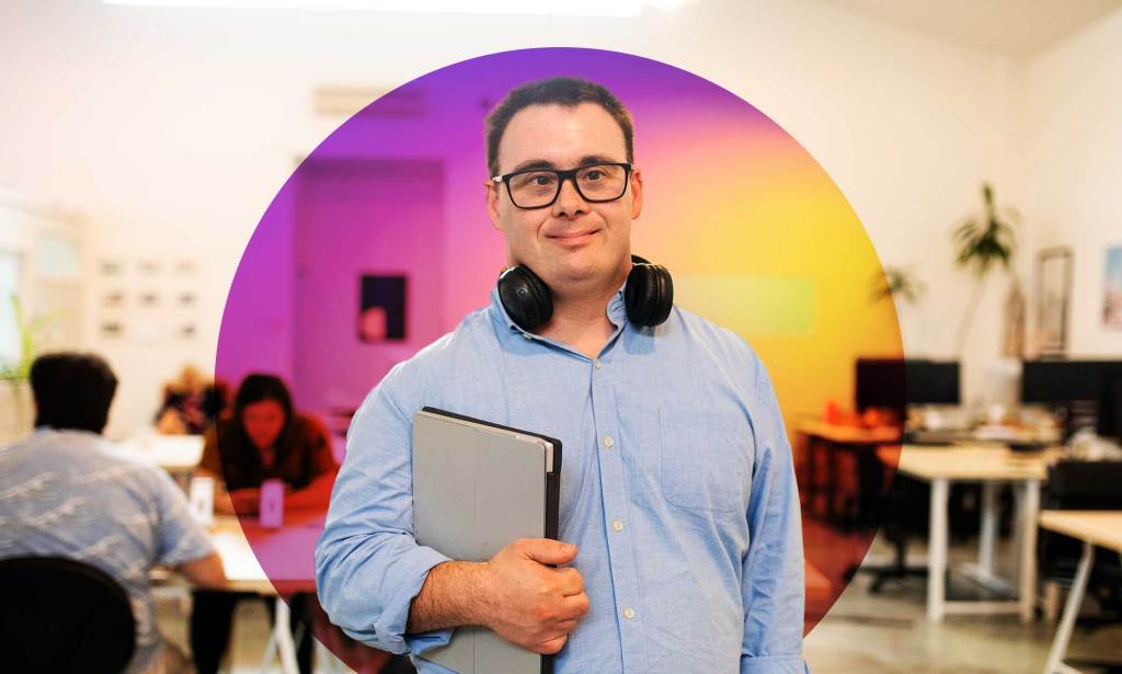 This is an image of a an with Down's Syndrome looking directly into the camera. He is in an office environment. He is wearing a blue shirt, dark eye glasses and a light blue buttoned shirt.