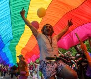 This is an image of a Pride parade. There is a man on a bicycle with his arms in the air and celebrating. He is under a massive Pride flag.