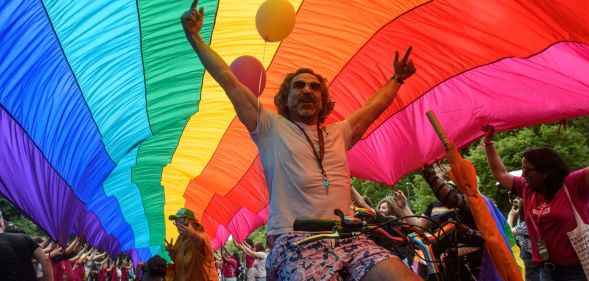 This is an image of a Pride parade. There is a man on a bicycle with his arms in the air and celebrating. He is under a massive Pride flag.