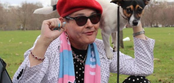 Trans activist Sarah Jane Baker, wearing a red cap and pink and blue scarf, holds a dog on her shoulder and raises her fist in solidarity with trans rights.