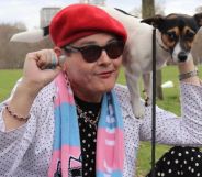 Trans activist Sarah Jane Baker, wearing a red cap and pink and blue scarf, holds a dog on her shoulder and raises her fist in solidarity with trans rights.
