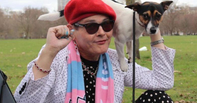 Trans activist Sarah Jane Baker, wearing a red cap and pink and blue scarf, holds a dog on her shoulder and raises her fist in solidarity with trans rights.