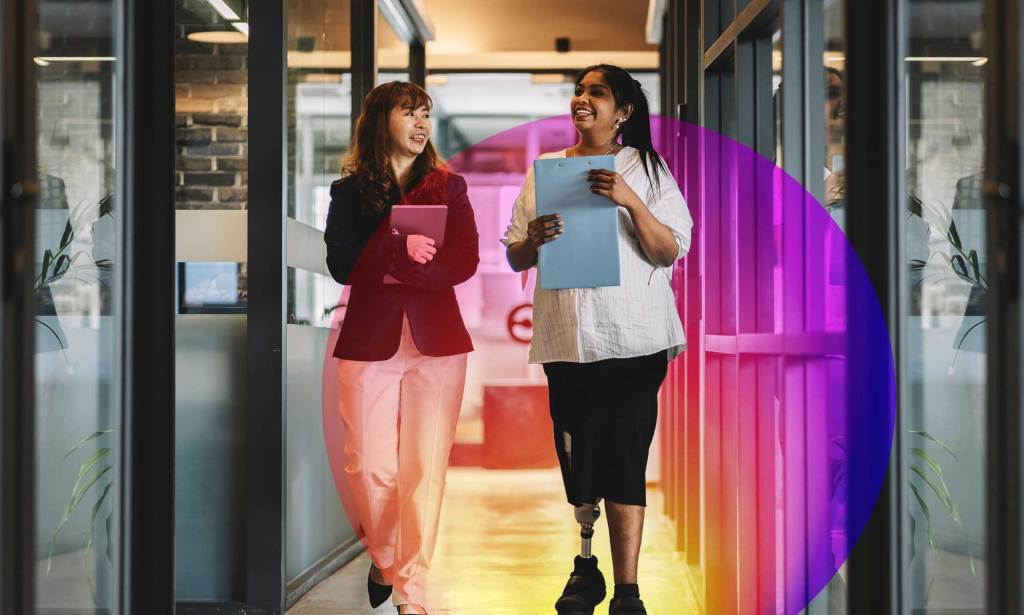 This is an image of two women walking down a hallway. The woman on the right is wearing a white top, dark trousers and a prosthetic leg. The woman on the left is wearing a dark top and light trousers.