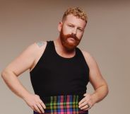 Non-binary, trans singer Tom Rasmussen wears a black vest and tartan shorts against a beige background.