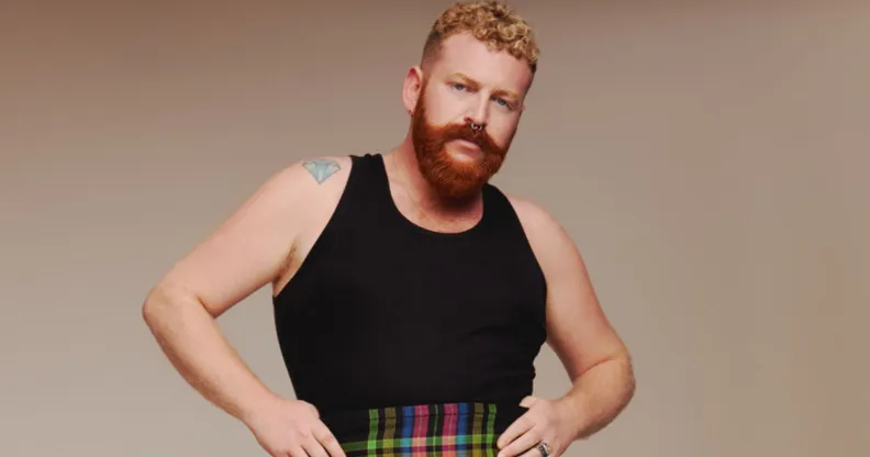 Non-binary, trans singer Tom Rasmussen wears a black vest and tartan shorts against a beige background.