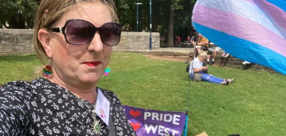 A member of Trans Pride South West taking a selfie in a park next to a trans flag waving in the air.