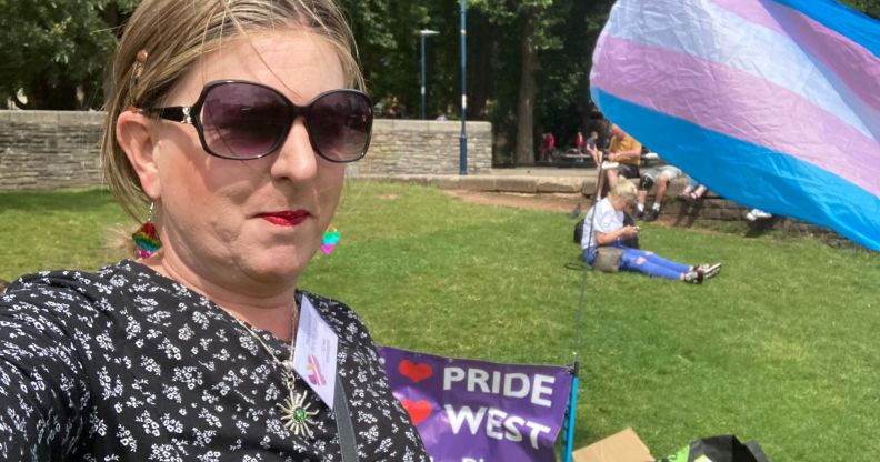 A member of Trans Pride South West taking a selfie in a park next to a trans flag waving in the air.