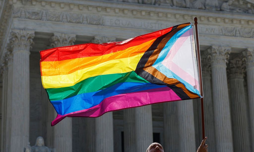 An individual holds an LGBTQ+ progress flag infront of the Supreme Court.