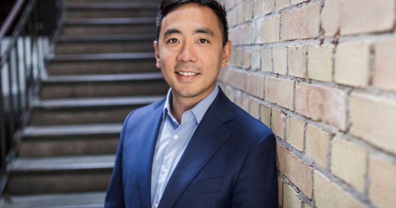 This is an image of Wayne Ting, the ceo of Lime. He is leaning on a brick wall. He is wearing a dark blue blazer with a light blue shirt He has black hair.
