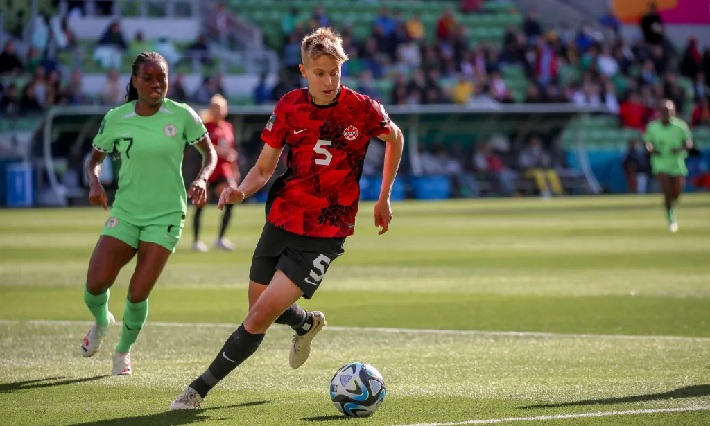 Canadian footballer Quinn wears a red and black uniform as they chase a football along the field at the 2023 Women's World Cup