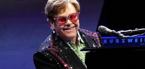 Singer Elton John wears a green shirt, dark jacket with sequins and red tinted sunglasses with gems along the rim as he plays the piano on stage