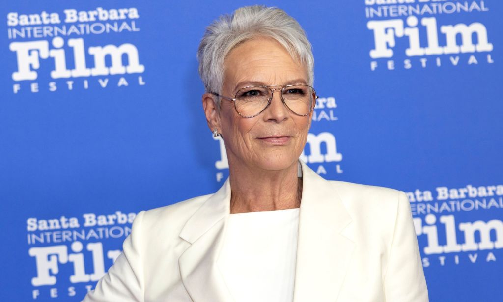 Actor Jamie Lee Curtis wears a white shirt and matching jacket as she poses in front of a blue background