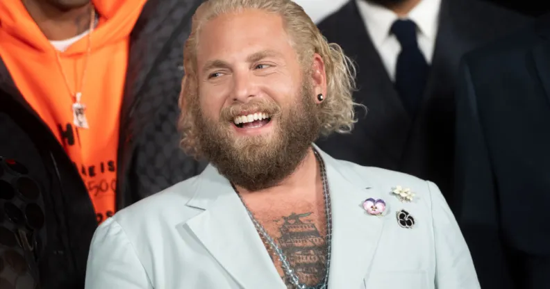 Jonah Hill, a white man with long bleached hair, wearing a light blue suit reveaing his tattooed chest
