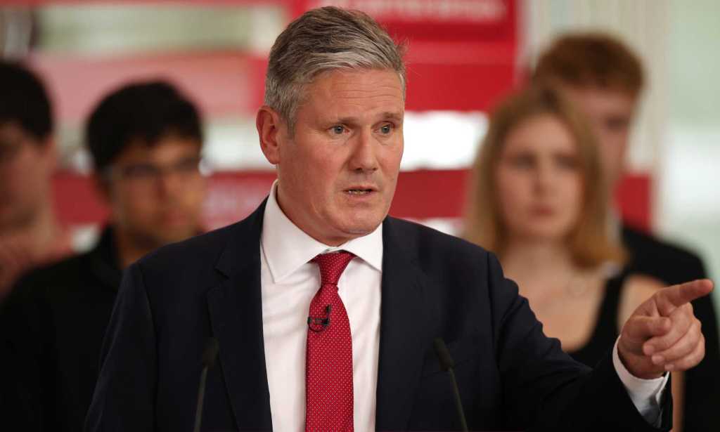 Keir Starmer, leader of the Labour Party, pictured speaking at a podium at a Labour event. He is wearing a black suit with a white shirt and red tie and he is gesturing to his left.
