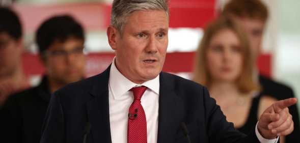 Keir Starmer, leader of the Labour Party, pictured speaking at a podium at a Labour event. He is wearing a black suit with a white shirt and red tie and he is gesturing to his left.