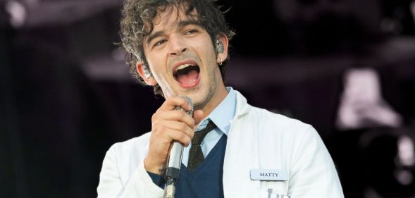 Matty Healy of The 1975 performing on stage wearing a lab coat, a sweater and a shirt and tie.