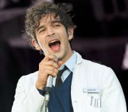 Matty Healy of The 1975 performing on stage wearing a lab coat, a sweater and a shirt and tie.