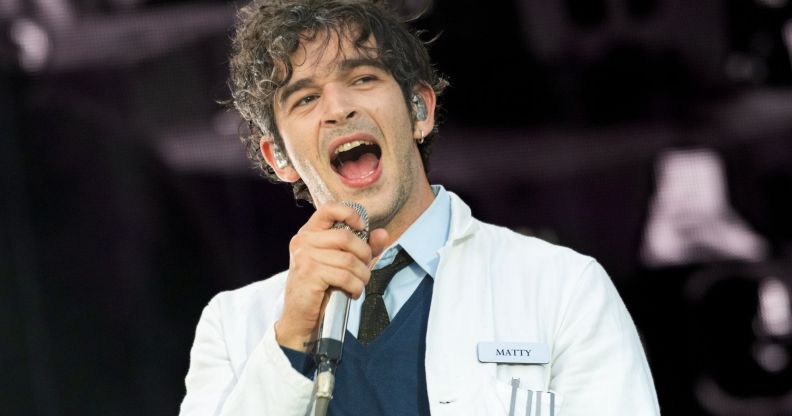 Matty Healy of The 1975 performing on stage wearing a lab coat, a sweater and a shirt and tie.