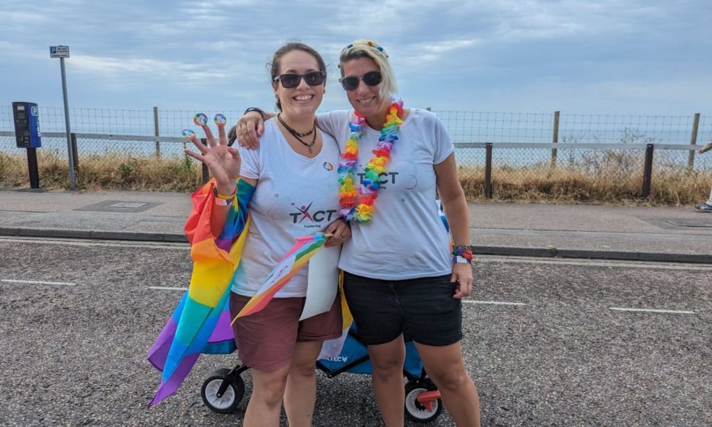 LGBTQ+ foster carers Nat and Terri stand side-by-side as they smile for the camera during a Pride March as they wear shirts with the logo of TACT