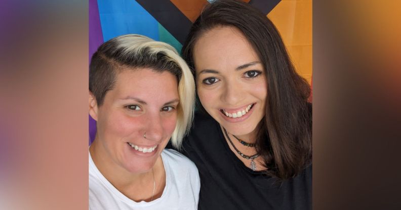 LGBTQ+ foster carers Nat and Terri stand side-by-side as they smile for the camera in front of a Progressive Pride flag