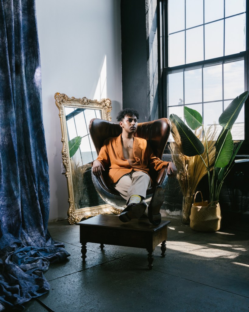 Rufus Sivaroshan, a music artist, pictured in a posed image sitting on an armchair with a mirror behind them and a decorated room in the background.