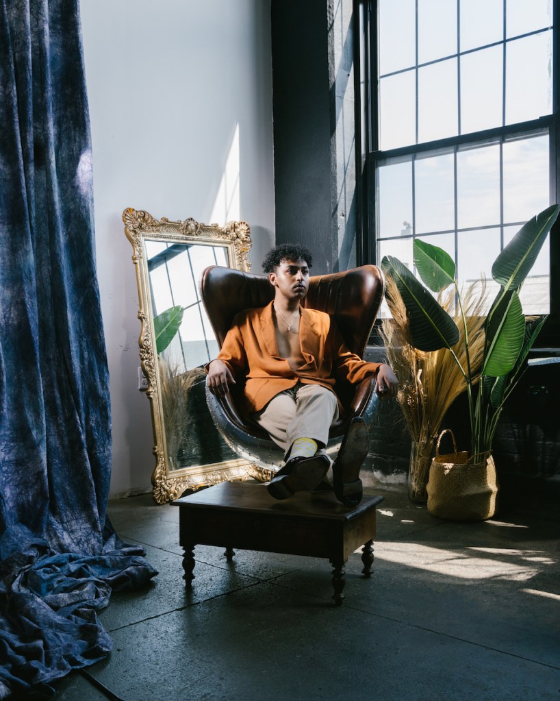 Rufus Sivaroshan, a music artist, pictured in a posed image sitting on an armchair with a mirror behind them and a decorated room in the background.