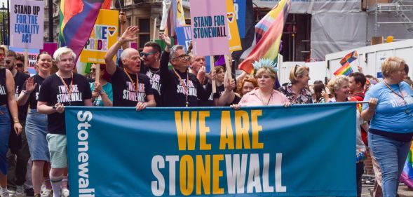 A group of people from LGBTQ+ charity Stonewall stand in a line next to each other as they walk through a Pride march holding up a sign reading 'We are Stonewall' stylised so the message 'We are one' can also be seen. In the lineup is Stonewall leader Nancy Kelley