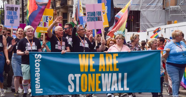 A group of people from LGBTQ+ charity Stonewall stand in a line next to each other as they walk through a Pride march holding up a sign reading 'We are Stonewall' stylised so the message 'We are one' can also be seen. In the lineup is Stonewall leader Nancy Kelley