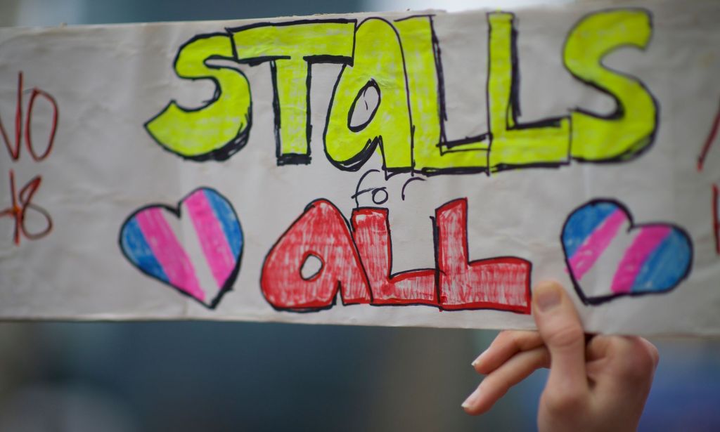 A person holds up a sign reading "stalls for all" during a demonstration for the LGBTQ+ and trans communities against trans bathroom bans and anti-trans legislation