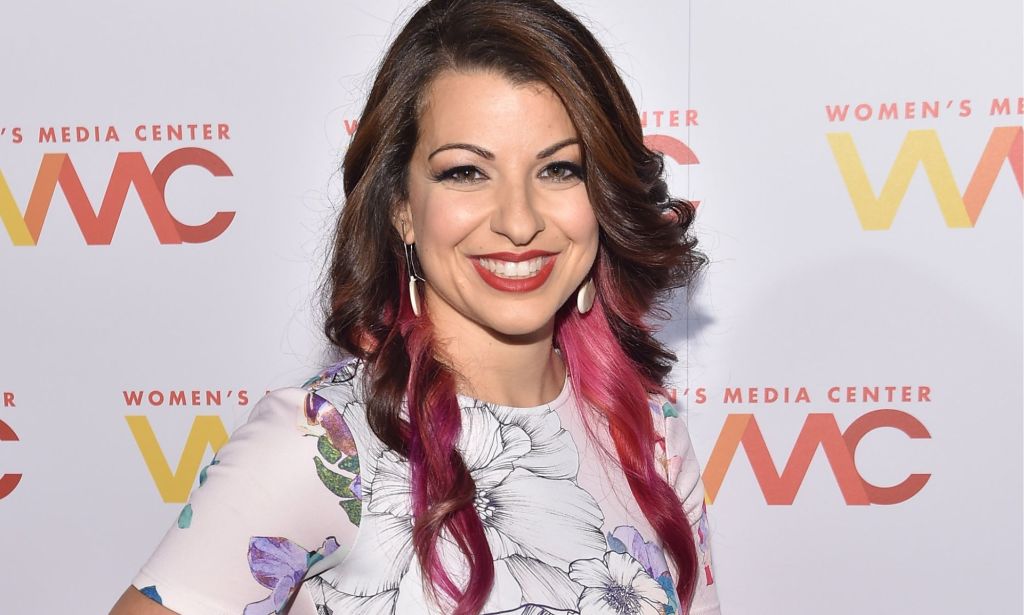 Anita Sarkeesian, wearing a white dress, smiles during a red carpet event.