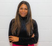 This is an image of Gabby Austen-Browne. She is standing on a white wall and is wearhing a black turtleneck. She has long brown hair and has her arms folded.