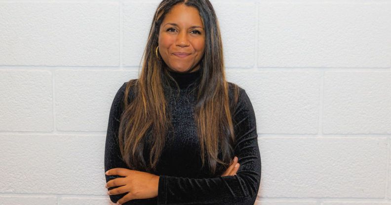 This is an image of Gabby Austen-Browne. She is standing on a white wall and is wearhing a black turtleneck. She has long brown hair and has her arms folded.