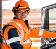 This is an image of a trans woman and HS2 worker, Kat, sitting inside a truck. She is wearing an orange hi-vis jacket and helmet. The door of the cab is open and she has one hand on the steering wheel.