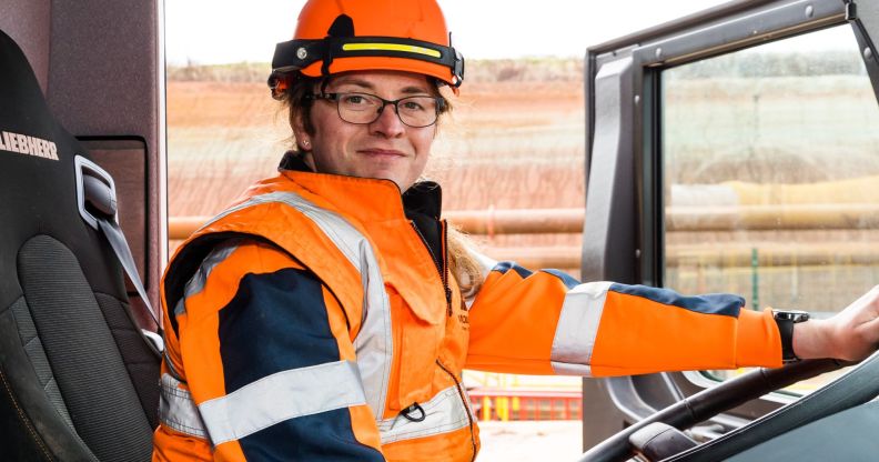 This is an image of a trans woman and HS2 worker, Kat, sitting inside a truck. She is wearing an orange hi-vis jacket and helmet. The door of the cab is open and she has one hand on the steering wheel.