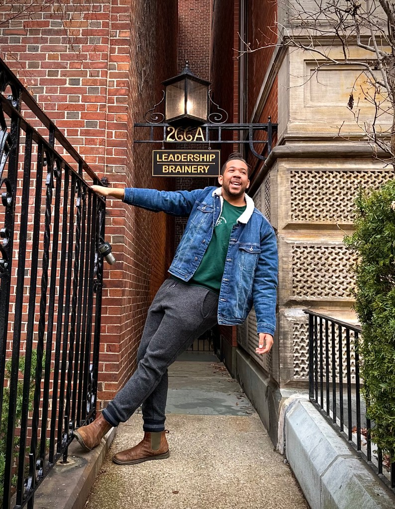 This is an image of JAKE Small. He is wearing a denim jacket and grey pants. He is standing on a pathway and one arm is hanging onto an iron railing.
