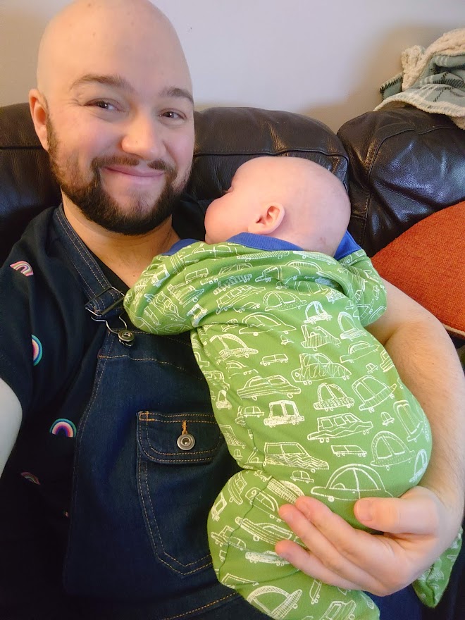 Jacob Stokoe pictured with his baby lying on his chest. The picture is a selfie which shows Jacob smiling as his baby, wearing a green baby grow, lies looking up at him.